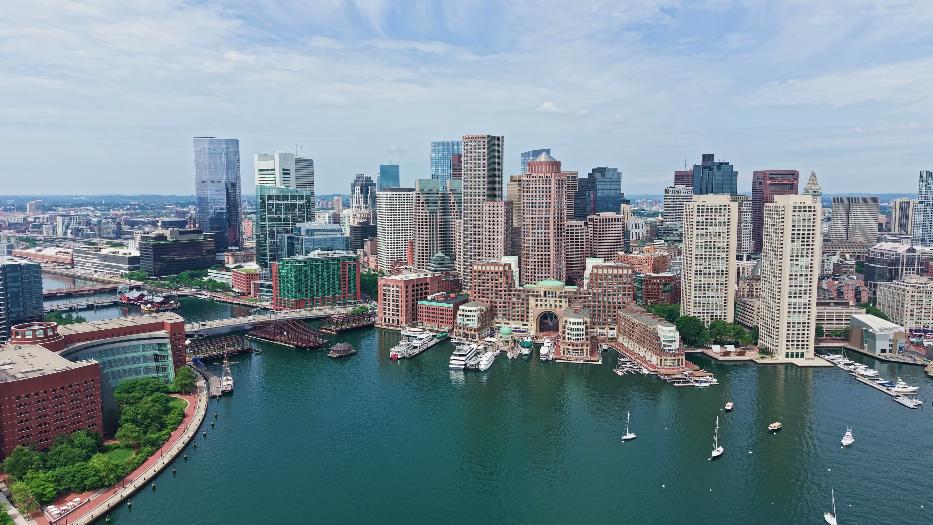 Aerial view of Boston's downtown skyline and waterfront across the harbor on a clear day, with sailboats on the water